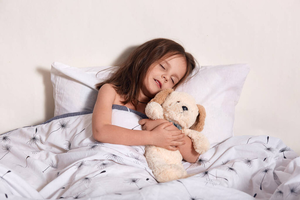 Οριζόντια προσωπογραφία του μικρού γλυκού χαριτωμένου παιδιού ξαπλωμένη στο κρεβάτι της, να κλείνει τα μάτια, να κοιμάται ήσυχα, να βλέπει όνειρα, να αγκαλιάζει το αρκουδάκι της, να έχει ευχάριστη έκφραση προσώπου. Έννοια παιδιών και ονείρων. - Φωτογραφία, εικόνα