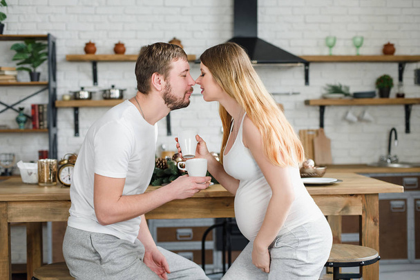 Pregnant Woman And Husband Having Breakfast at Kitchen drink coffee or fresh juice and kissing. - Фото, зображення