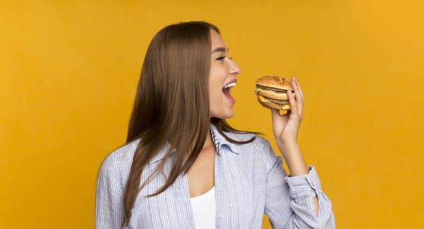 Chica feliz comiendo hamburguesa malsana de pie sobre fondo amarillo, Panorama
 - Foto, Imagen