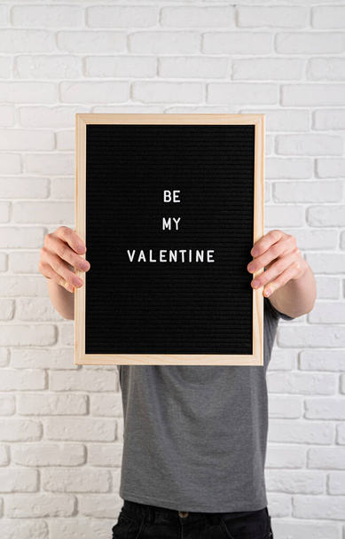 Mężczyzna trzymający czarną tablicę z filcem ze słowami Be My Valentine przeciwko białej ceglanej ścianie - Zdjęcie, obraz