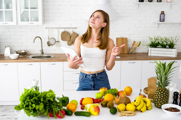 A young girl in the kitchen with a tablet in her hands is looking for recipes for cooking. A table with a variety of vegetables and fruits for a healthy vegetarian diet. - Foto, Imagen