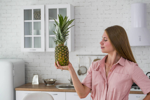 A beautiful young girl looks at a big pineapple that is holding in her hands. White kitchen on the background. - 写真・画像