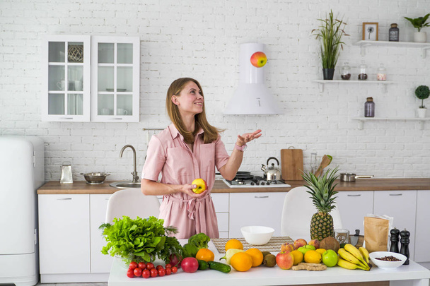 Cheerful girl in the kitchen near the table with fruits and vegetables throws an apple. Healthy vegetarian food. - Foto, Bild