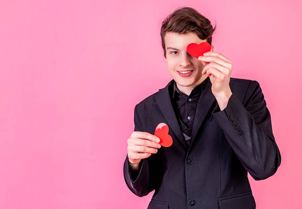 Young smiling man holding heart shaped valentine cards in front of his eyes on pink background with copy space - Photo, Image