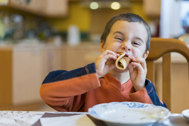 Boldog kisfiú palacsintát eszik. Portré kis aranyos kisfiú gyermek ül az asztalnál otthon eszik palacsintát. - Fotó, kép