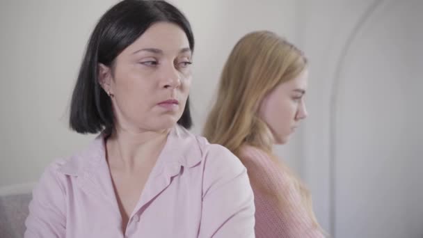 Close-up portrait of brunette Caucasian woman sitting back to back with blond girl. Portrait of mother argued with daughter at home. Family relationship, conflict, problems. - Záběry, video