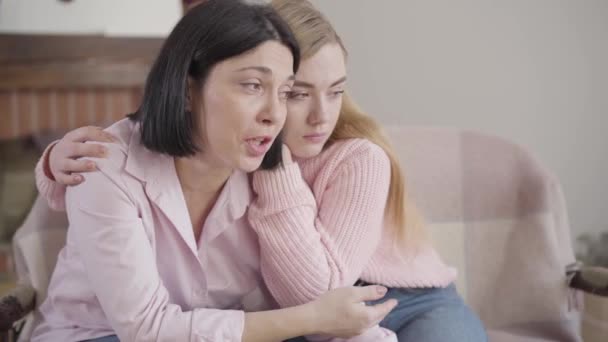 Close-up side view of brunette Caucasian woman sitting with blond girl on couch and talking. Teenage daughter calming down mother at home. Family problems, unity, support. - Felvétel, videó