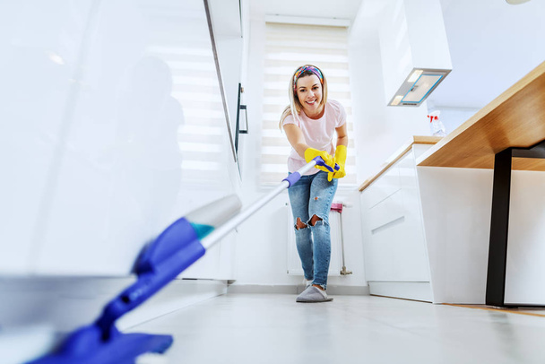 Smiling Caucasian blond housewife with rubber gloves on cleaning kitchen floor with mop. Weekly chores are getting done. - Fotoğraf, Görsel