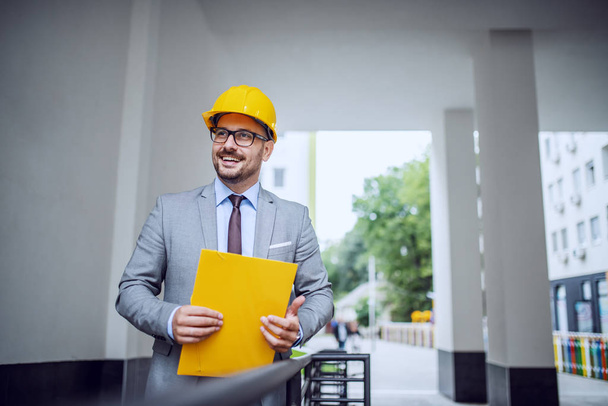 Handsome smiling elegant architect in suit and with helmet on head standing outdoors with folder in hands. - Photo, Image