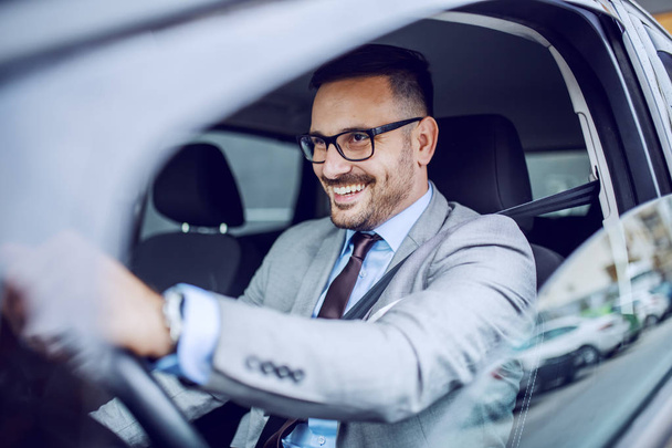 Caucasiano atraente sorrindo elegante empresário sem barba em terno e óculos dirigindo seu carro caro
. - Foto, Imagem