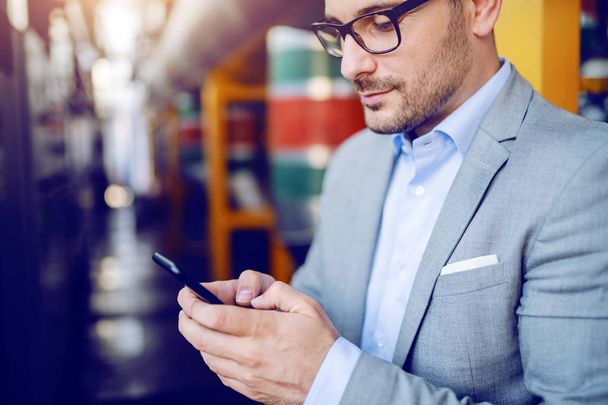 Bělošský podnikatel v šedém obleku a s brýlemi stojící v továrně a pomocí chytrého telefonu. - Fotografie, Obrázek