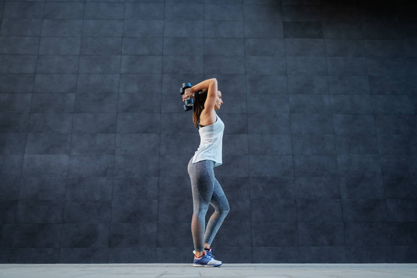 Vista lateral de la deportista en forma de pie al aire libre con pesas detrás de las espaldas y haciendo ejercicios para las espaldas
. - Foto, imagen