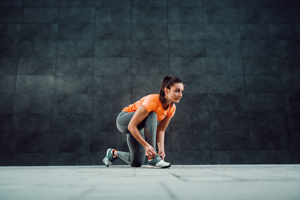 Fit attractive caucasian brunette in sportswear and with ponytail kneeling and tying shoelace before running. - Foto, afbeelding