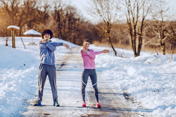 Lächelndes kaukasisches Paar in Sportkleidung und mit gesunden Gewohnheiten, das die Arme ausstreckt, während es auf der Landstraße steht. Winterzeit. Fitness in der Natur. - Foto, Bild