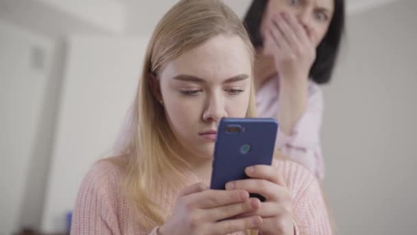 Portrait of blurred Caucasian woman looking at smartphone screen over girls shoulder with shocked facial expression. Worried parent spying teen daughter. Reliance, relationship. - Footage, Video