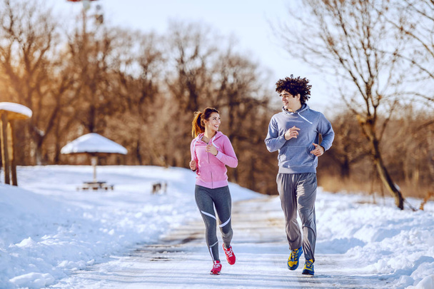Happy smiling caucasian couple in sportswear running in nature while looking at each other. Winter time. - Photo, Image