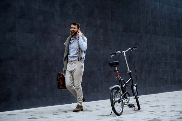 Gleeful guapo hombre de negocios caucásico de moda escuchando música, cantando y sosteniendo maletín mientras va a trabajar. Junto a él está la bicicleta. En el fondo es la pared gris
. - Foto, imagen