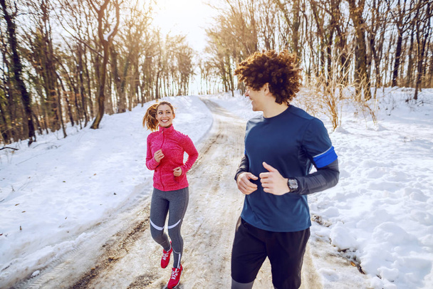 Χαρούμενο χαμογελαστό ζευγάρι Καυκάσιων με αθλητικά να τρέχουν στη φύση κοιταζόμενοι. Χειμώνας. - Φωτογραφία, εικόνα