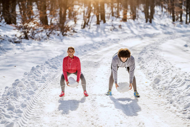 glücklich fittes kaukasisches Paar in Sportkleidung, das auf einem Fußweg hockt und Ausdauer mit großen Schneebällen übt. Winterzeit. Outdoor-Fitnesskonzept. - Foto, Bild