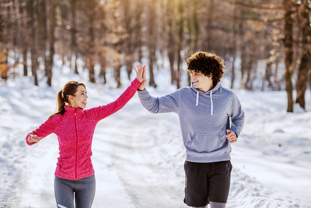 Cheerful caucasian handsome couple in sportswear running on outdoor and giving high five to each other. Outdoor fitness concept. Wintertime. - Photo, Image