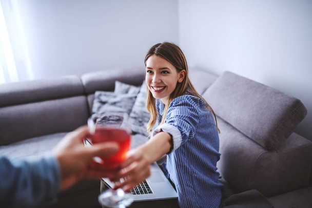 Young smiling charming caucasian brunette in stripped shirt sitting on sofa in living room and getting glass of wine from her boyfriend. - Photo, Image