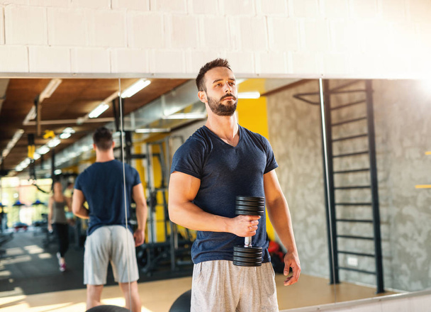 Musculoso hombre serio en ropa deportiva sosteniendo mancuerna. En el fondo su reflejo en el espejo
. - Foto, imagen