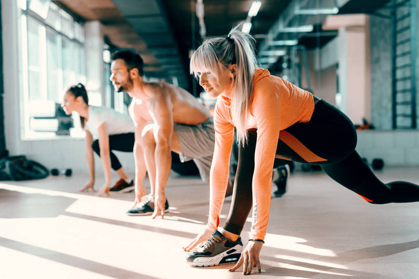 Small group of people with healthy habits doing stretching exercises on a gym floor. Selective focus on blonde woman. In background mirror. - Photo, Image