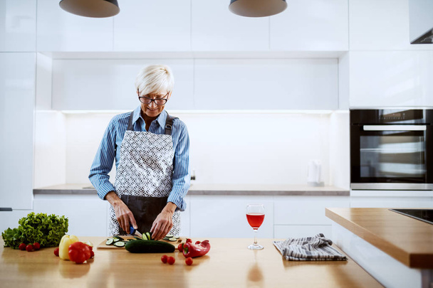 Χαμογελώντας λευκή ηλικιωμένη γυναίκα με ποδιά στέκεται στην κουζίνα και κόβει αγγούρι. Στον πάγκο της κουζίνας είναι πιπεριές, μαρούλι και ένα ποτήρι κρασί. - Φωτογραφία, εικόνα