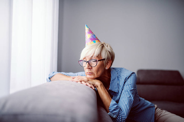 Droevige blanke blonde senior vrouw met verjaardagspet op het hoofd zittend op de bank in de woonkamer en leunend. - Foto, afbeelding