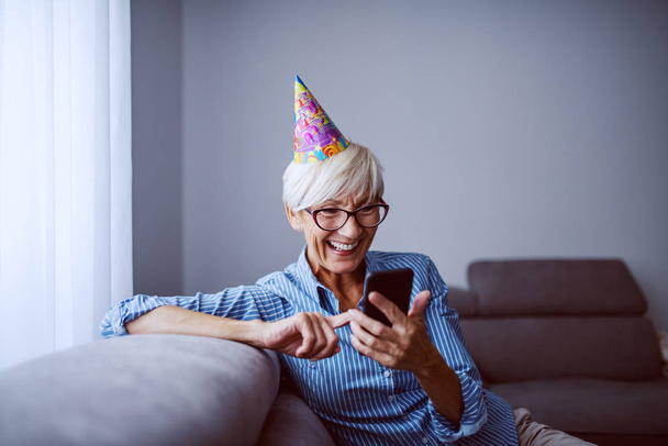 Overjoyed smiling caucasian blond senior woman with birthday cap on head sitting on sofa in living room and reading birthday messages on smart phone. - Photo, Image