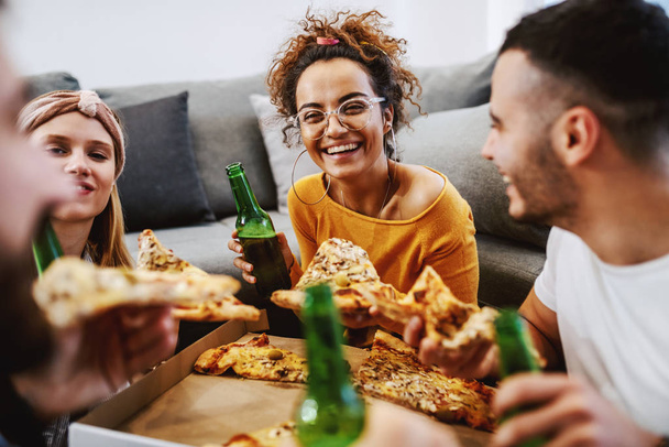 Група друзів, які сидять на підлозі у вітальні, п'ють пиво та їдять піцу. Вечірка вдома
. - Фото, зображення