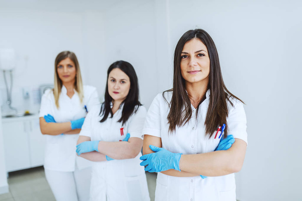 Portrait of beautiful Caucasian female lab assistant in white uniform and with rubber gloves standing with arms crossed. In background are her colleagues. - Photo, Image