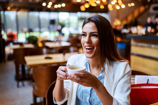 charmante kaukasische lachende Brünette sitzt in der Cafeteria, hält eine Tasse frischen Kaffee in der Hand und genießt die freie Zeit. - Foto, Bild