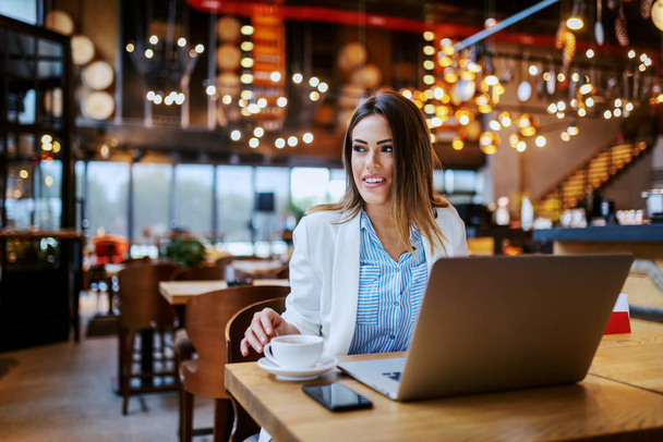 Bájos mosolygós kaukázusi barna nő ül a kávézóban, és készül dolgozni online. Az asztalon laptoppal és friss kávéval. - Fotó, kép