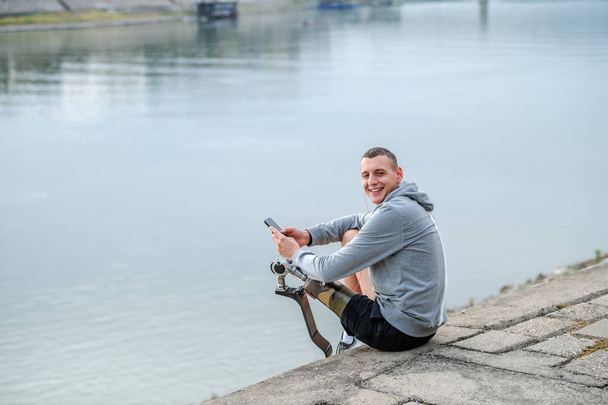 Веселий білий спортсмен з інвалідністю у спортивному одязі та зі штучною ногою, що сидить поруч з річкою та засуджує смартфон, дивлячись на камеру
. - Фото, зображення