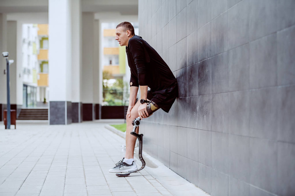 Longitud completa del guapo deportista caucásico con pierna artificial apoyada en la pared y descansando de correr
. - Foto, Imagen
