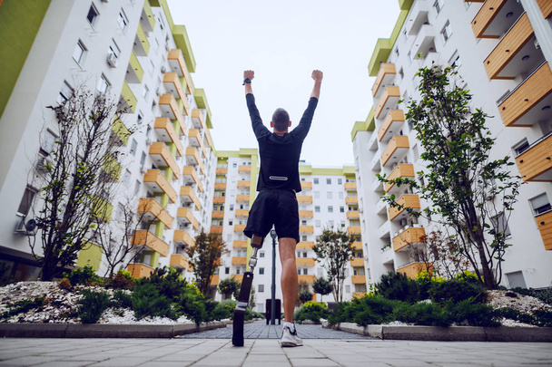 Vue arrière du beau sportif avec jambe artificielle debout avec les mains en l'air à l'extérieur entouré de bâtiments
. - Photo, image