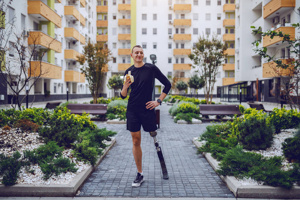 A vonzó kaukázusi sportoló teljes hossza mesterséges lábbal áll a parkban épületekkel körülvéve és banánt eszik.. - Fotó, kép