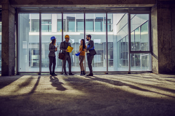 Τέσσερις αφοσιωμένοι εργατικοί αρχιτέκτονες στέκονται δίπλα σε ένα μεγάλο παράθυρο στο παλιό κτίριο και μιλάνε για το πώς σκοπεύουν να το ξαναχτίσουν για να γίνουν σύγχρονοι.. - Φωτογραφία, εικόνα