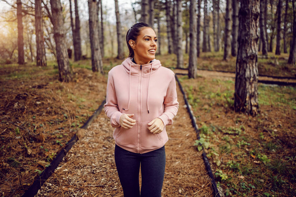 Smiling young woman in sportswear running in woods. Healthy lifestyle. Running in nature concept. - Photo, Image