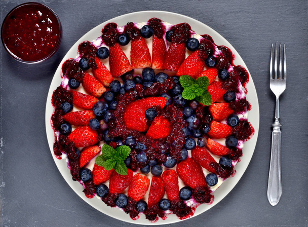 Délicieux gâteau fraises fraîches, bleuets, confiture de cerises et feuilles de menthe sur plaque d'ardoise. Couteau et bol avec confiture à l'arrière-plan. Vue du dessus
. - Photo, image