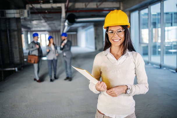 Jovem bem sucedido arquiteto feminino caucasiano atraente com capacete na cabeça de pé dentro do edifício em processo de construção e segurando a papelada nas mãos
. - Foto, Imagem