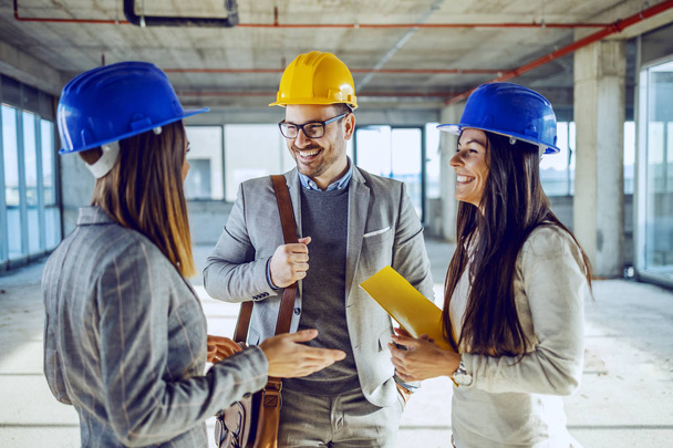 Group of young highly motivates attractive caucasian architects with helmets on heads standing in building in construction process and having casual talk on pause. - Photo, Image