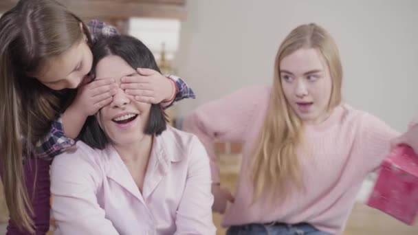 Two Caucasian daughters giving surprise present to their adorable mother. Happy brunette woman receiving gift at home. Happiness, leisure, lifestyle. - Footage, Video