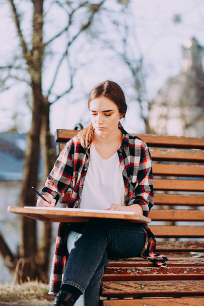 Young woman artist draws a picture while sitting on a bench. Painter artist holds a wooden tablet for painting and a brush in his hand - Foto, Imagem