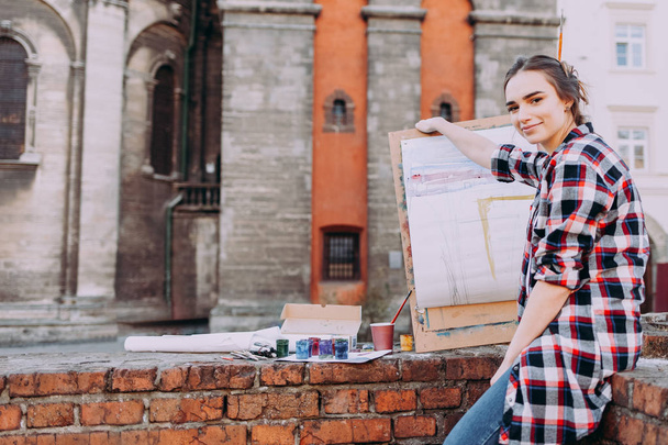 Woman artist paints a picture while sitting on a brick wall on a background of old architecture. Girl spends leisure drawing pictures on a city street. Smiling female artist - Photo, Image