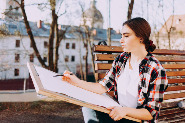 La joven artista dibuja un cuadro mientras está sentada en un banco. Artista pintor sostiene una tableta de madera para pintar y un pincel en su mano
 - Foto, imagen