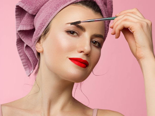 Close-up of a beautiful woman with natural make-up and clean skin with a towel on her head. Girl combing thick eyebrows and looking at the camera. Cosmetology and spa. Morning time and make up. Red li - Фото, изображение