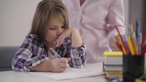 Close-up portrait of tired Caucasian girl sitting at the table and writing. Unrecognizable woman shaking hands at the background. Mom scolding daughter for problems in education. - Footage, Video
