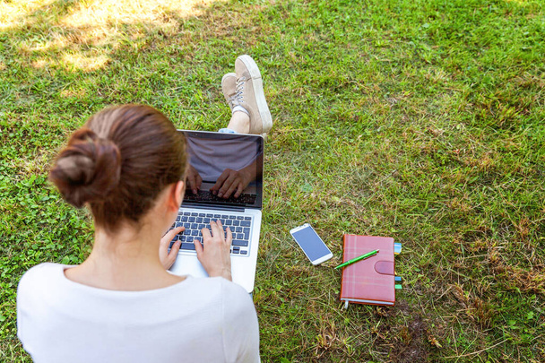 Young woman sitting on green grass lawn in city park working on laptop pc computer. Freelance business concept - Fotografie, Obrázek